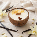 Bougie Luminante Vanille dans son contenant en noix de coco, entourée de gousses de vanille et de fleurs