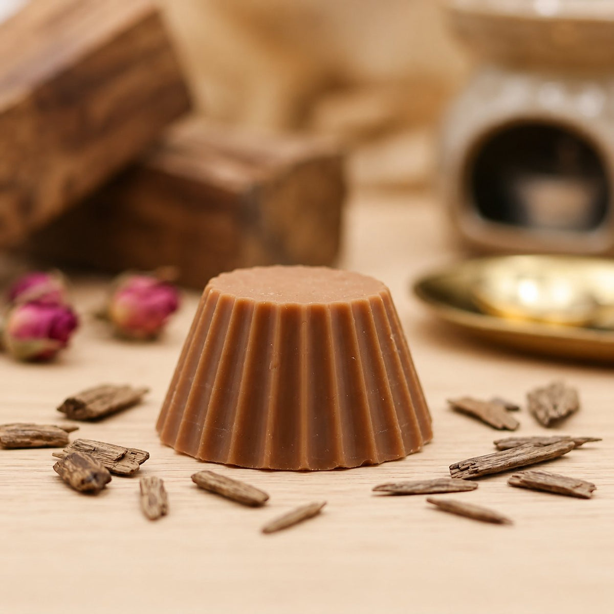 Fondant parfumé Luminante Bois de Oud entouré d'éclats de bois, devant un brûle-parfum en verre