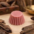 Fondant parfumé Luminante Cuir et bois vu de près, entouré de morceaux de bois brut, devant une coupelle en laiton