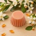 Fondant parfumé Luminante Fleur d'oranger, pastille de cire rosée posée sur un plan en bois clair, entourée de fleurs d'oranger blanches et de pétales orangés