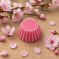 Fondant parfumé Luminante Fleur de Cerisier, pastille de cire rose posée sur un plan en bois clair, entourée de fleurs et de pétales de cerisier