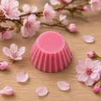 Fondant parfumé Luminante Fleur de Cerisier, pastille de cire rose posée sur un plan en bois clair, entourée de fleurs et de pétales de cerisier