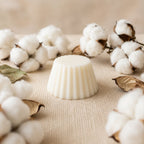 Fondant parfumé Luminante Fleur de Coton sur une toile écrue, cerné de fleurs de coton et de feuilles séchées