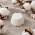 Fondant parfumé Luminante Fleur de Coton, pastille de cire blanche posée sur une toile de lin froissée, entourée de branches de coton