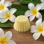Fondant parfumé Luminante Fleur de Tiaré sur une table en bois, entouré de fleurs de frangipanier et de feuilles vertes