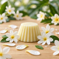 Fondant parfumé Luminante Fleur de Tiaré, pastille de cire crème posée sur un plan en bois clair, entourée de fleurs blanches à cœur jaune et de gouttes d'eau