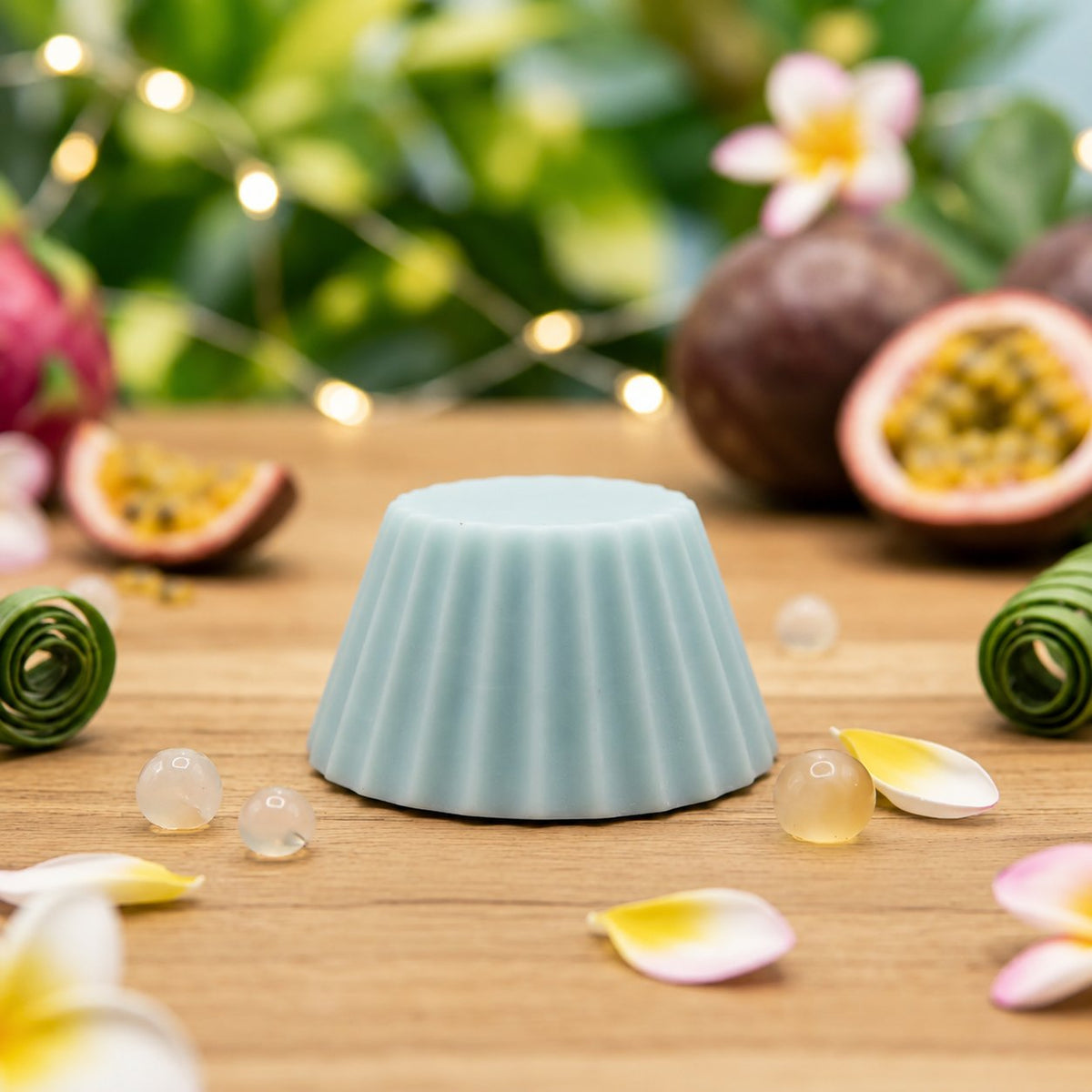 Fondant parfumé Luminante Fruits Exotiques, pastille de cire bleutée posée sur un plan en bois, entre des fruits de la passion et des fleurs de frangipanier