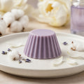 Fondant parfumé Luminante Lotus blanc coton, pastille de cire mauve sur une assiette en céramique, entourée de fleurs de coton, de brins séchés et de pétales blancs