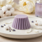 Fondant parfumé Luminante Lotus blanc coton, pastille de cire mauve sur une assiette en céramique, entourée de fleurs de coton, de brins séchés et de pétales blancs