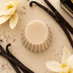 Fondant parfumé Luminante Vanille vu de dessus sur un fond crème, entre des gousses de vanille et des fleurs d'orchidée crème