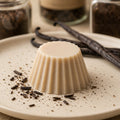 Fondant parfumé Luminante Vanille, pastille de cire beige sur une assiette en grès, entre des gousses de vanille et des éclats de gousse