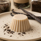 Fondant parfumé Luminante Vanille, pastille de cire beige sur une assiette en grès, entre des gousses de vanille et des éclats de gousse