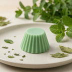 Fondant parfumé Luminante Verveine Menthe, pastille de cire verte sur une assiette blanche, entre des feuilles de menthe fraîche et des feuilles séchées