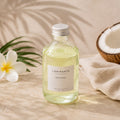 Recharge Luminante Tiaré-Coco, flacon en verre entre des fleurs de tiaré et une noix de coco ouverte