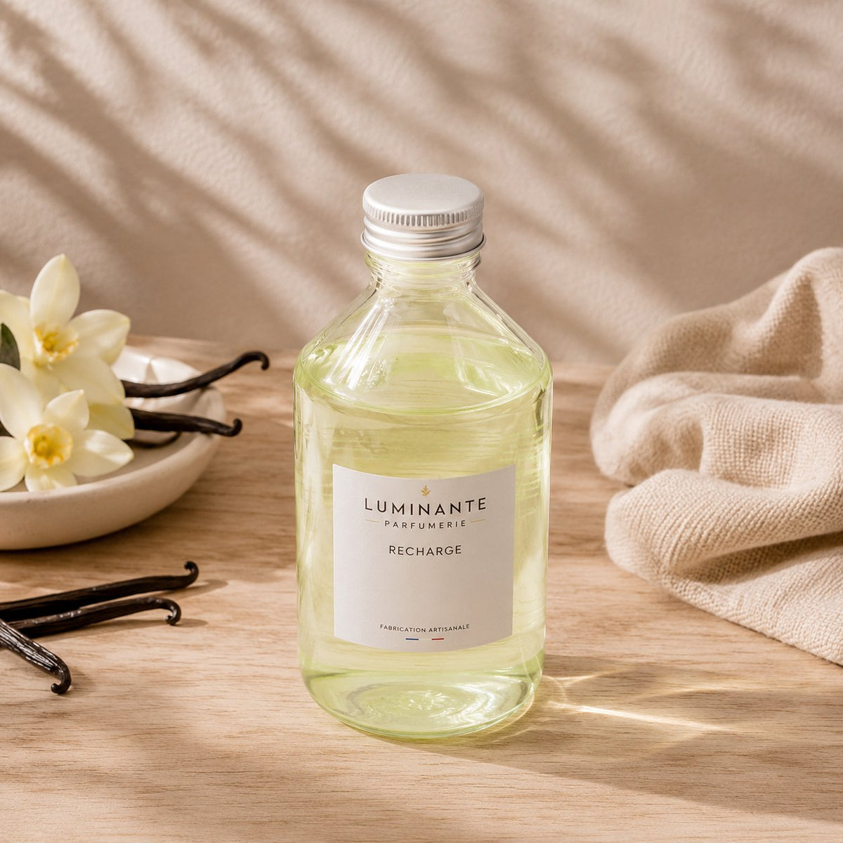 Recharge Luminante Vanille-Musc, flacon en verre entre des gousses de vanille et des fleurs crème